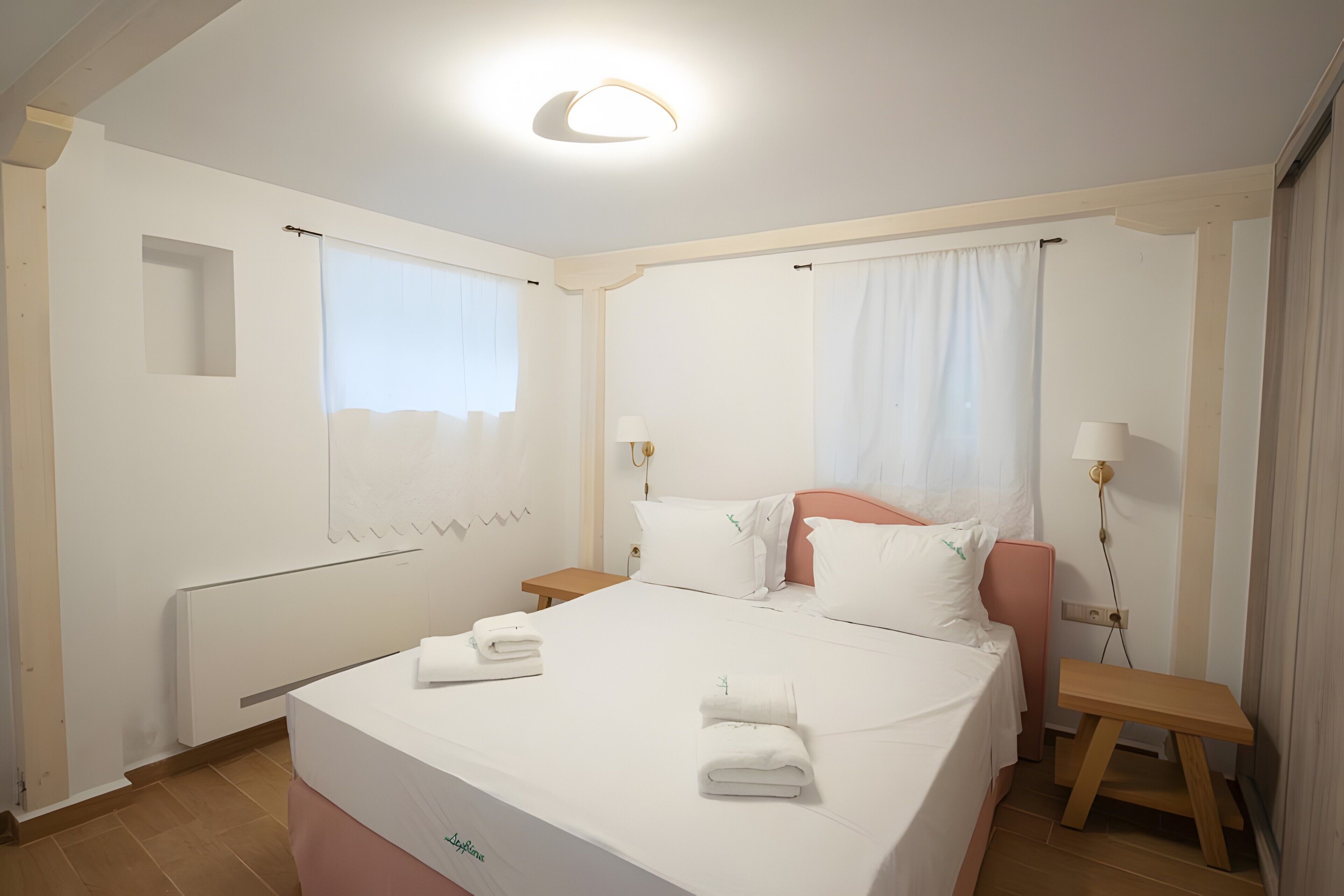 A hotel room with a bed, a bedside table, a nightstand, a window, and a wall-mounted lamp.