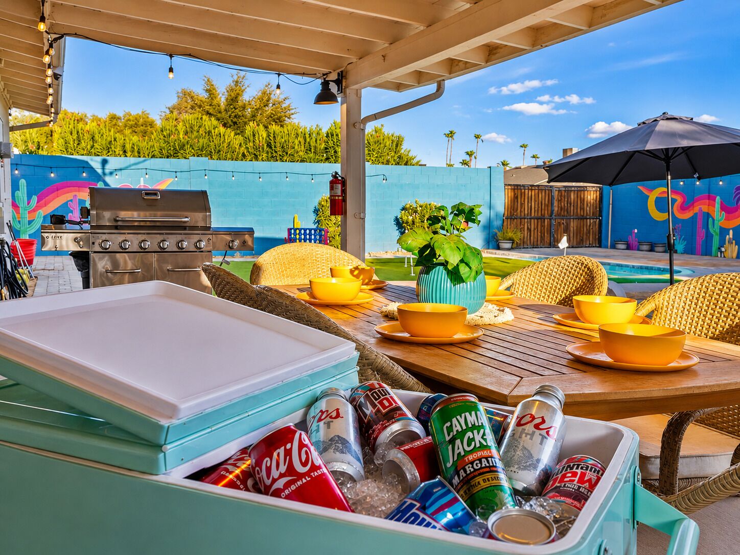 Une terrasse extérieure équipée d’un barbecue, d’une table à manger, de chaises et d’une glacière remplie de boissons.