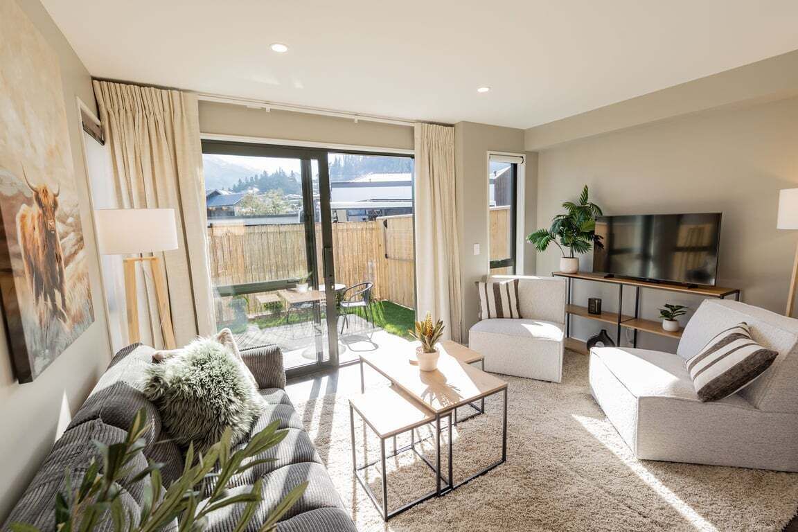 A modern living room with a sofa, coffee table, and TV. There is a sliding glass door leading to a balcony with a view of the outdoors.