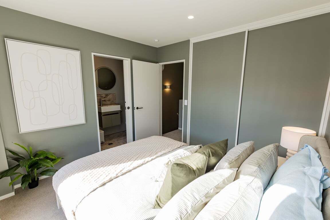 Ein Schlafzimmer mit einem Bett, Kissen, einer Nachttischlampe, einer Topfpflanze und einer Tür zu einem Badezimmer.