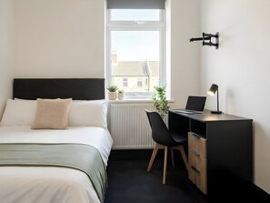 A hotel room with a bed, a desk with a laptop, a chair, a radiator, and a window with a view of buildings. - House Sleeps 6 with Patio (Cardiff)