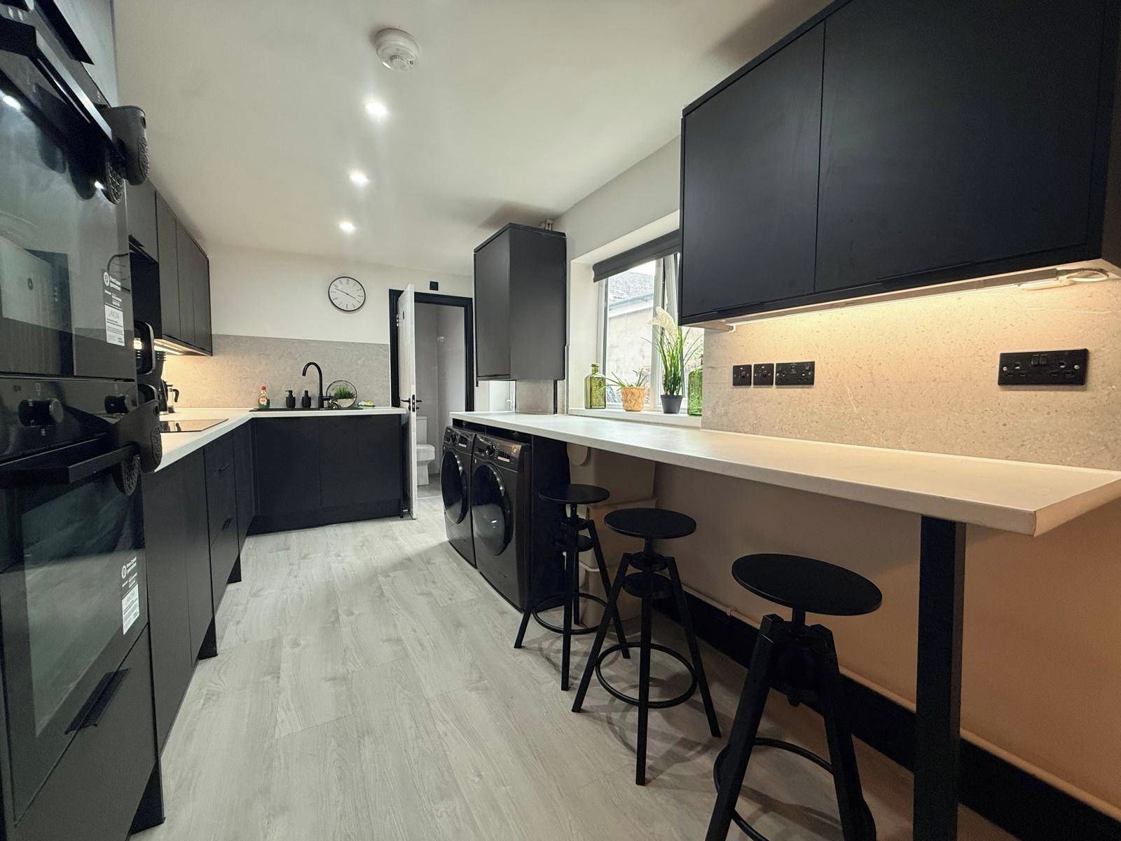 Une cuisine moderne avec des meubles de couleur sombre, un îlot central doté de tabourets et une horloge bien visible.