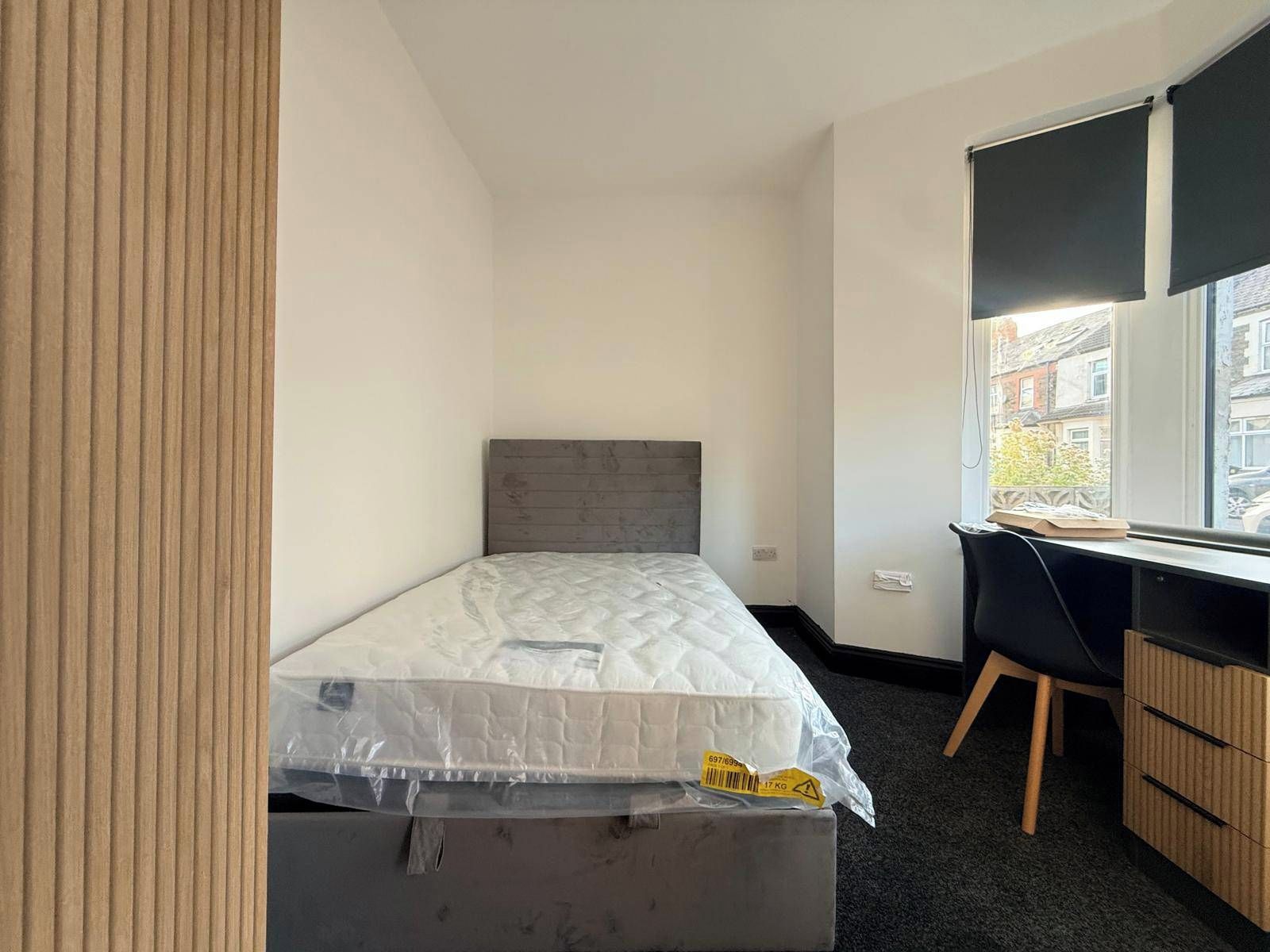 A small, single-bed room with a grey headboard, a desk with a chair, and a window with blinds.