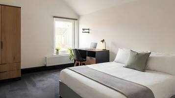 A bedroom with a bed, a desk with a laptop, a radiator, and a window.