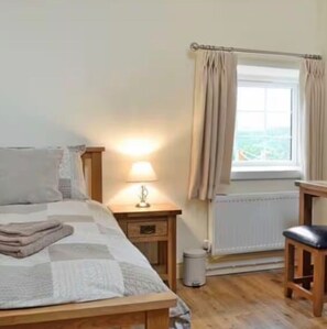A bedroom with a bed, bedside lamp, window with curtains, radiator, and a small stool. - Luxury Holiday cottages Sleeps 10 (Ystrad Meurig)