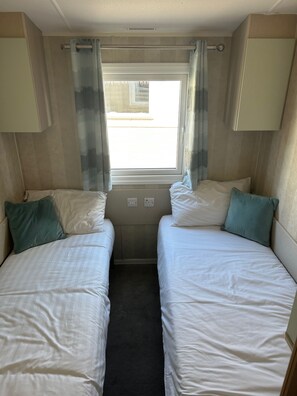 A compact room with two single beds, a window with curtains, and a small table. - Cabin Sleeps 8 with Sea Views and Parking (Morecambe)