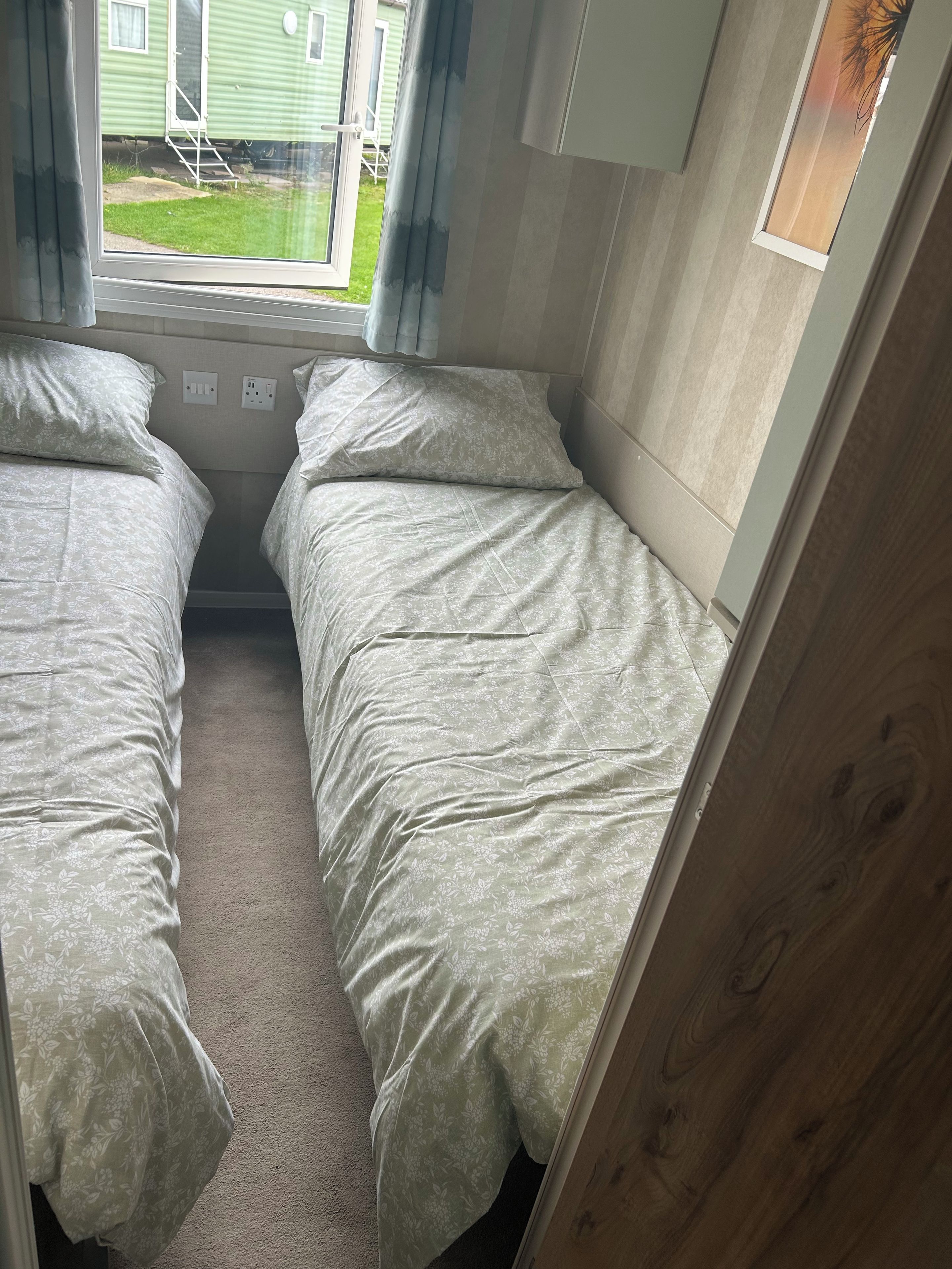 A bedroom with two beds, a window with curtains, and a view of a green house outside.