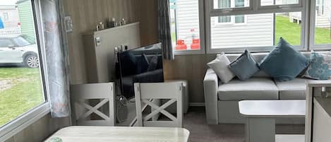 A compact room with a sofa, a television, a dining table, and a window with a view of a vehicle and a building outside.