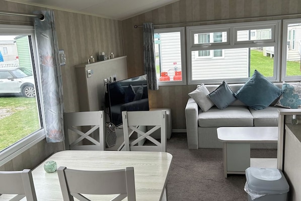 A compact room with a sofa, a television, a dining table, and a window with a view of a vehicle and a building outside.
