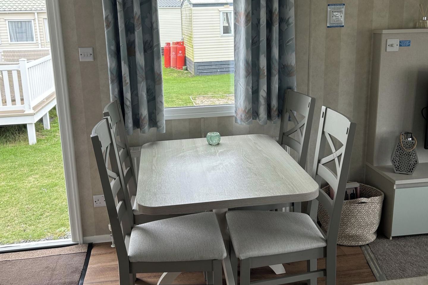 A small dining area with a table and chairs, a window with curtains, and a view of a grassy yard and a building outside.