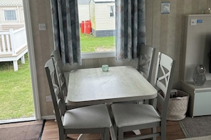 A small dining area with a table and chairs, a window with curtains, and a view of a grassy yard and a building outside.