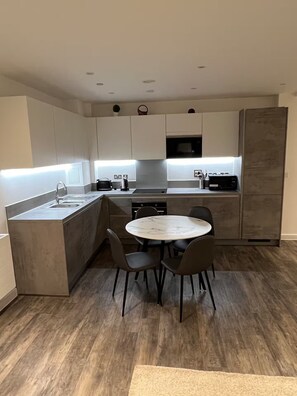 A modern kitchen with a central island, two chairs, a dining table, and a built-in oven. - Cosy Barking Apartment with City Views (Barking)