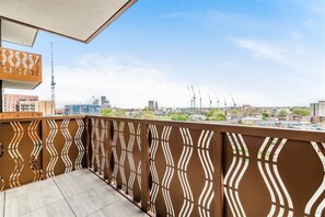 Ein Balkon mit schmiedeeisernem Geländer, der auf eine Stadtlandschaft mit Gebäuden und Kränen blickt.