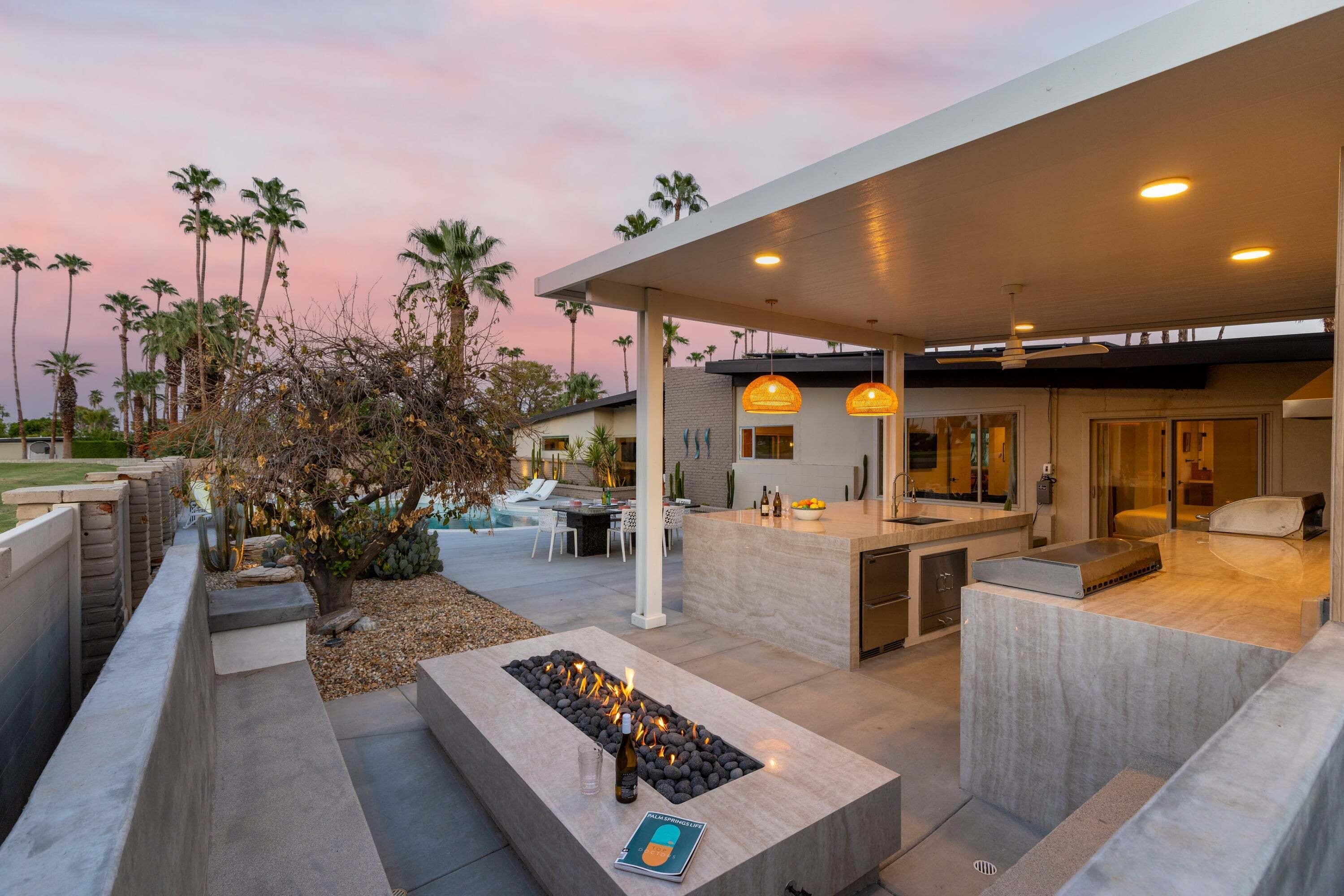 Un área de cocina exterior moderna con una chimenea integrada, asientos y una piscina al fondo.