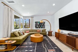 Ein modernes Wohnzimmer mit einer Eckcouch, einem runden Couchtisch, einem Fernseher und einem großen Fenster mit Vorhängen.