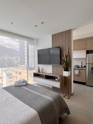 A modern bedroom with a large bed, a TV mounted on a wooden panel, and a view of the city through large windows. - Vrbo Property (Quito)