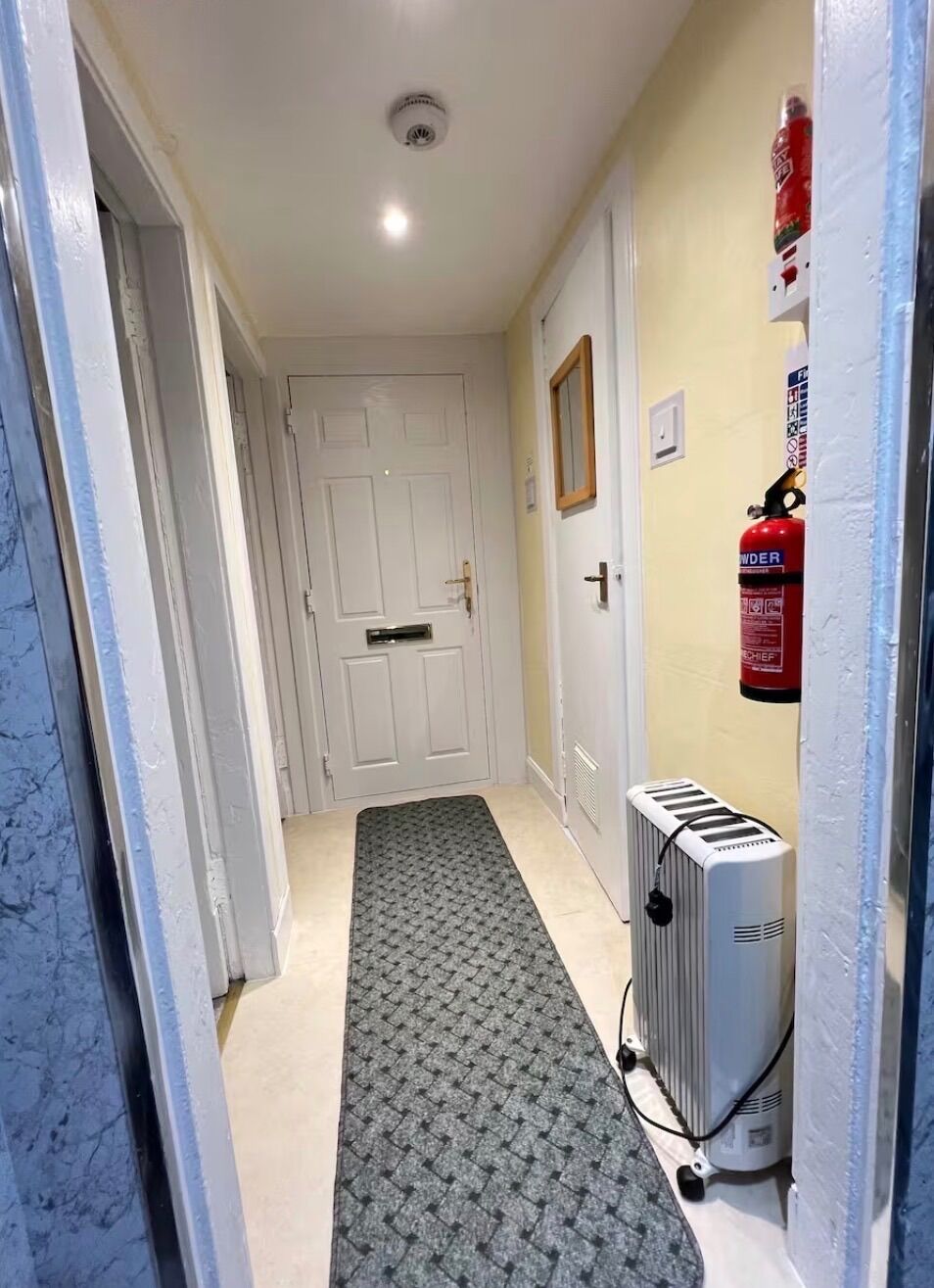Un couloir avec un radiateur, un extincteur et une porte au fond.