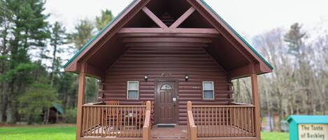 A wooden cabin with a front porch and a door, surrounded by a grassy area with trees.
