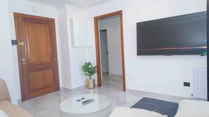 A modern living room with a wooden door, a potted plant, a round coffee table, and a flat-screen TV mounted on the wall.