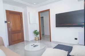 A modern living room with a wooden door, a potted plant, a round coffee table, and a flat-screen TV mounted on the wall.