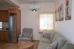 Uma sala de estar com sofá, poltrona estampada, mesa de centro de madeira e área de cozinha com geladeira.