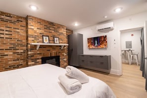 A modern hotel room with a brick fireplace, a flat-screen TV, a desk with a chair, and a neatly made bed. - Spruce Villa - 1st Floor Oversized King 1BR (Philadelphia)