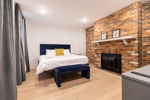 Ein modernes Schlafzimmer mit Kamin aus Ziegelsteinen, einem Bett mit weißer Bettwäsche, einer Bank und einem Schreibtisch mit einem Laptop.