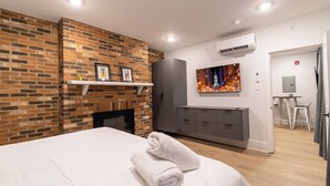 Una habitación de hotel moderna con pared de ladrillo decorativa, chimenea, cama grande con una toalla enrollada, armario gris, televisor montado en la pared y equipo de aire acondicionado.