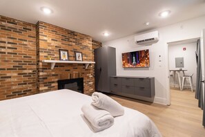 A modern hotel room with a brick accent wall, a fireplace, a large bed with a rolled towel, a grey wardrobe, a TV mounted on the wall, and an air conditioning unit. - Thena Hotel - Oversized 1 Bedroom (Philadelphia)