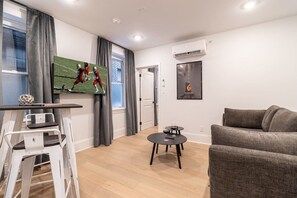 Ein modernes Wohnzimmer mit einem grauen Sofa, einem kleinen runden Tisch mit Weingläsern, einem Fernseher, der ein Sportspiel zeigt, und einer Wand-Klimaanlage.