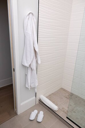Ein Badezimmer mit Duschkabine aus Glas, einem weißen Handtuch an der Wand und einem Paar weißer Hausschuhe auf dem Boden.