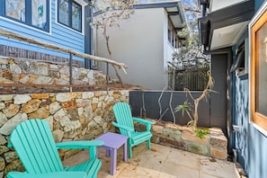 A patio with two green Adirondack chairs and a small table, a stone wall, and a tree.