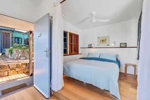 Une chambre à coucher avec un lit, un ventilateur de plafond, une fenêtre avec des volets en bois et un mur en pierre à l’extérieur.