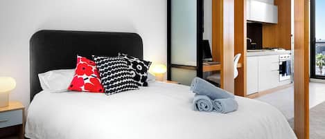 A hotel room with a bed, pillows, a nightstand, a lamp, a towel, and a view of a kitchen and balcony.