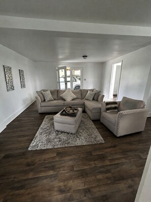 Ein modernes Wohnzimmer mit einem Ecksofa, einem Pouf und einem gemusterten Teppich.