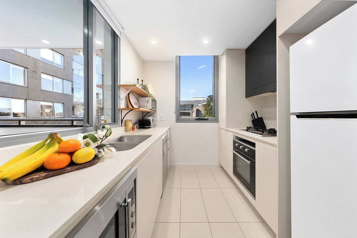 A modern kitchen with a large window, a built-in oven, and a countertop with fruits and a coffee maker.