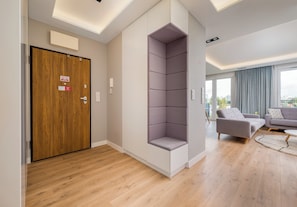 A modern living room with a wooden floor, a grey sofa, and a large window with a view of buildings. - Imielin Signature Apartment (Warszawa)