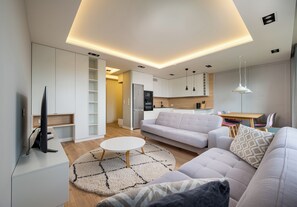 A modern living room with a sofa, a coffee table, and a TV. There is a kitchen area with cabinets and a refrigerator. - Imielin Signature Apartment (Warszawa)