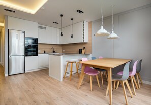 A modern kitchen with white cabinets, a stainless steel refrigerator, a built-in oven, and a wooden dining table with colorful chairs. - Imielin Signature Apartment (Warszawa)