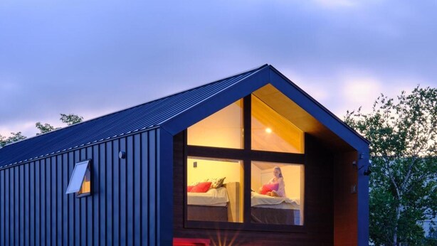 Um edifício moderno de dois andares com telhado inclinado, quarto visível com cama e janela, e uma porta vermelha.
