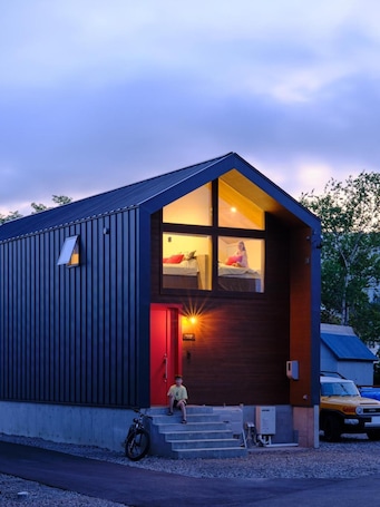 A modern two-story building with a sloped roof, a visible bedroom with a bed and window, and a red door.