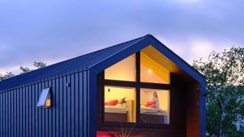 Ein modernes, zweistöckiges Gebäude mit geneigtem Dach, ein sichtbares Schlafzimmer mit Bett und Fenster sowie eine rote Tür.