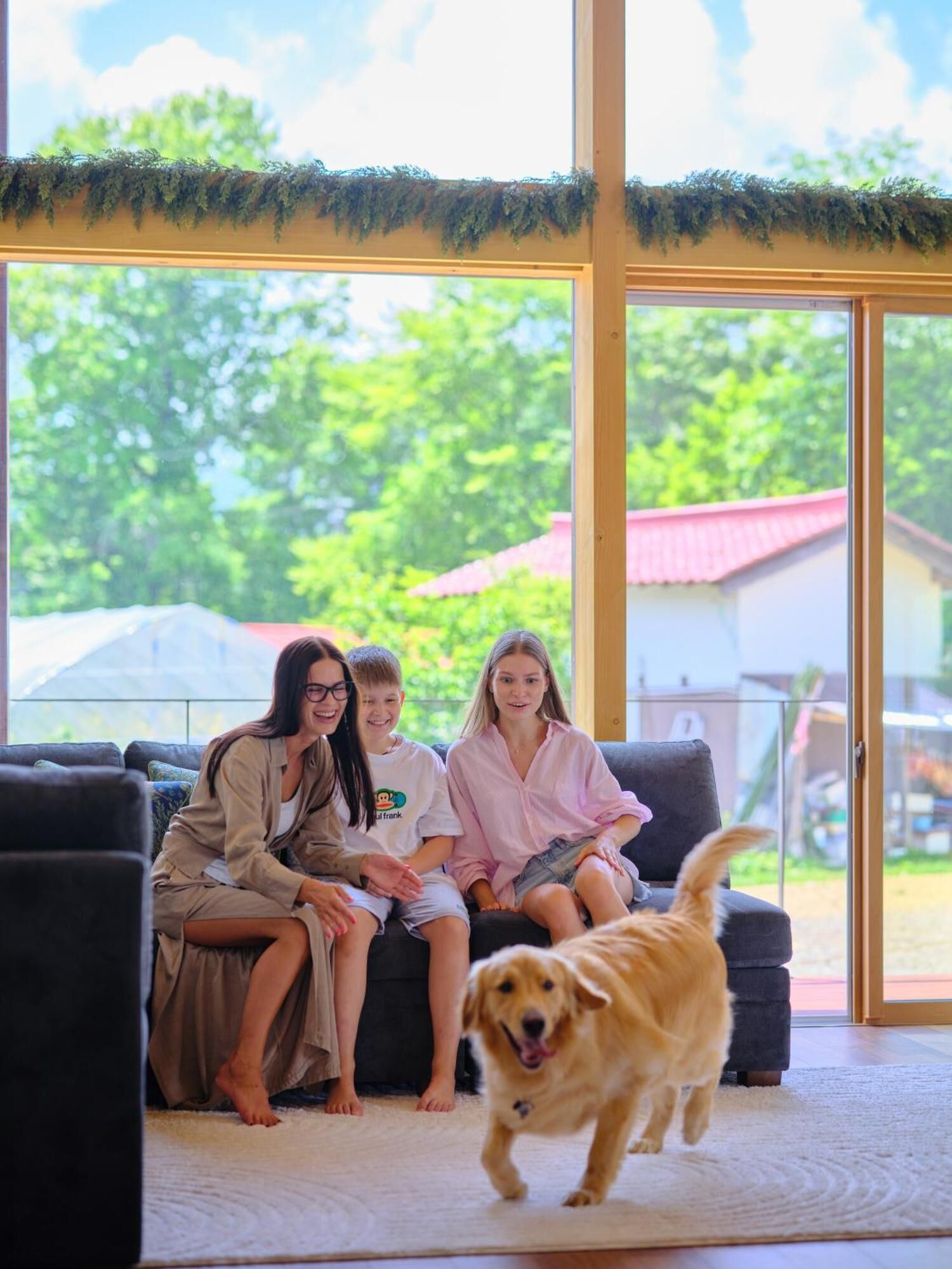 A group of people sitting on a couch in a room with large windows, a dog in the foreground, and a view of greenery outside.