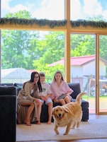 Un grupo de personas sentadas en un sofá en una habitación con ventanas amplias, un perro en primer plano y vistas a la vegetación al exterior.