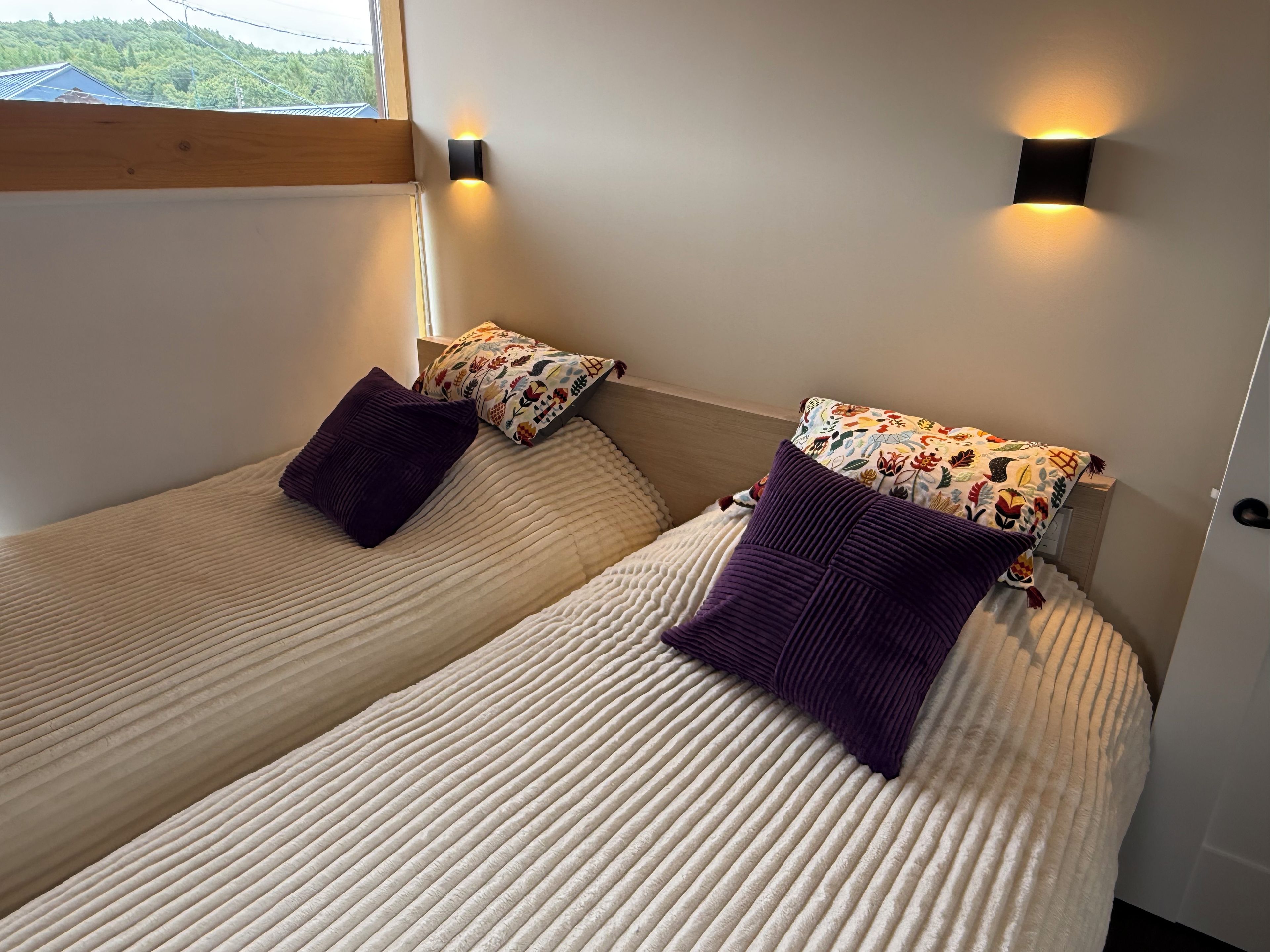 Deux lits simples avec des têtes de lit, une fenêtre donnant sur un espace verdoyant et des lampes fixées au mur.