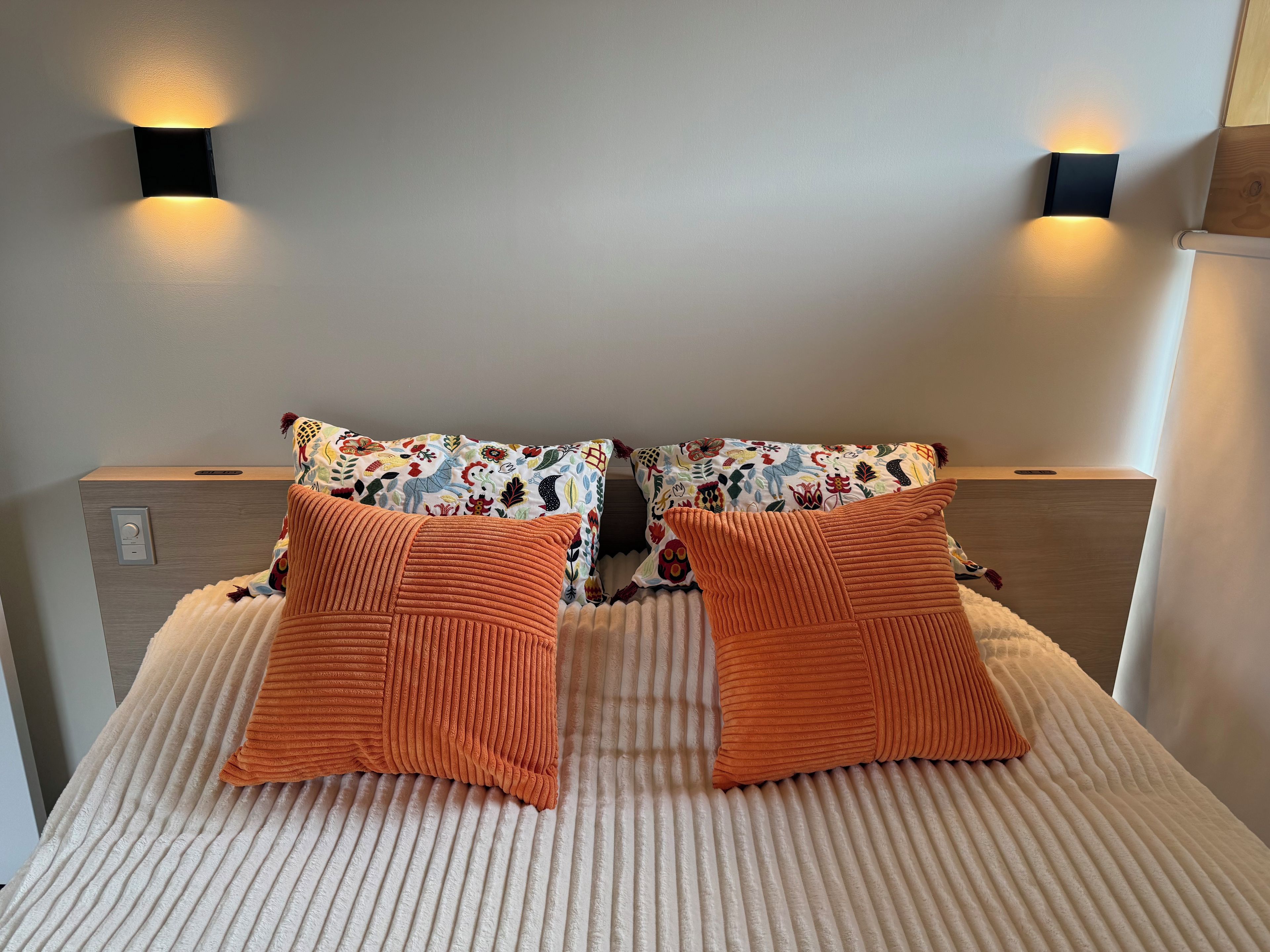 A bed with two orange pillows, a patterned pillow, and a headboard with a light switch.