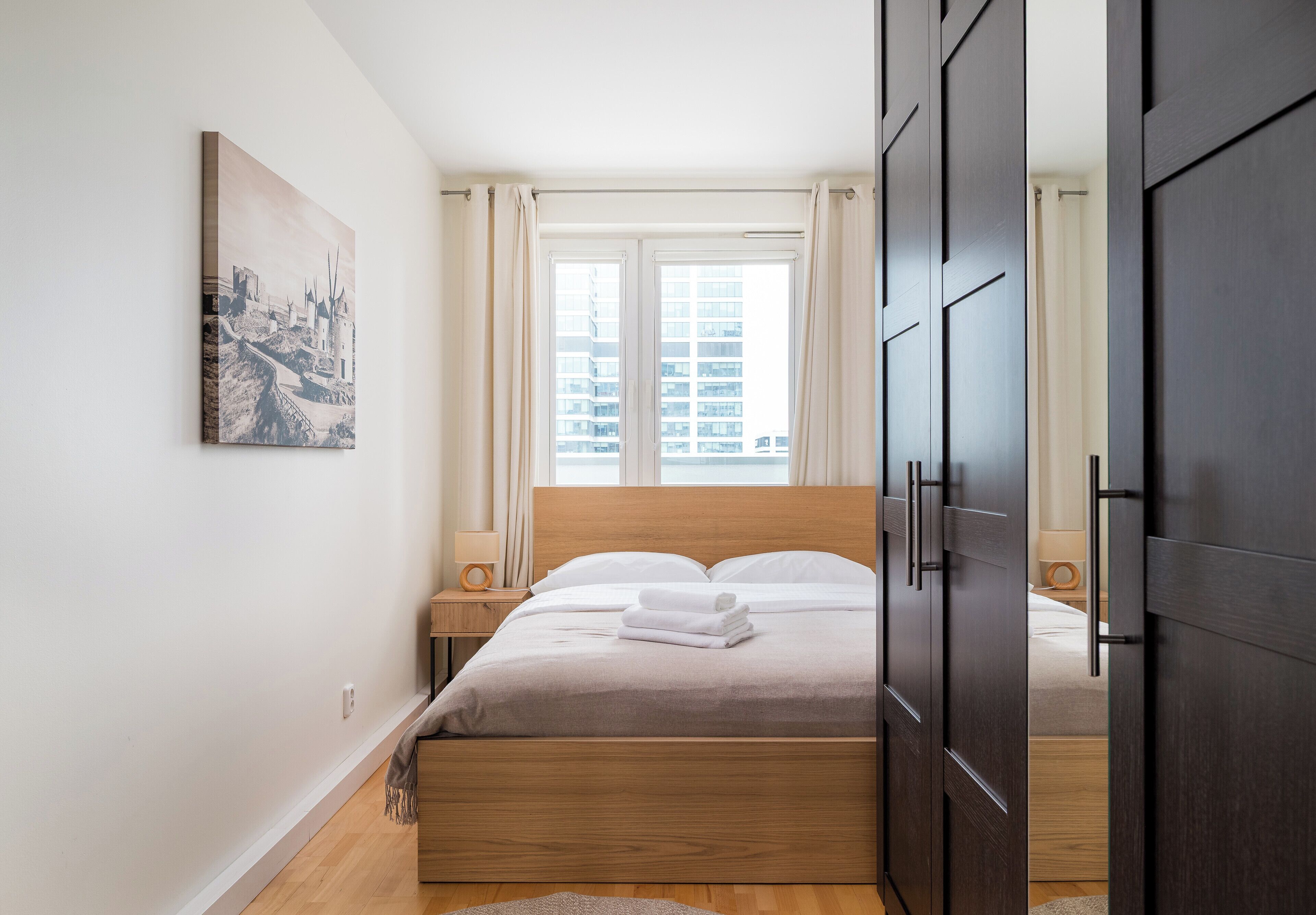 A bedroom with a wooden bed, a nightstand, a lamp, a painting on the wall, and a window with curtains.