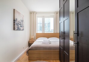 Ein Schlafzimmer mit einem hölzernen Bett, einem Nachttisch, einer Lampe, einem Bild an der Wand und einem Fenster mit Vorhängen.