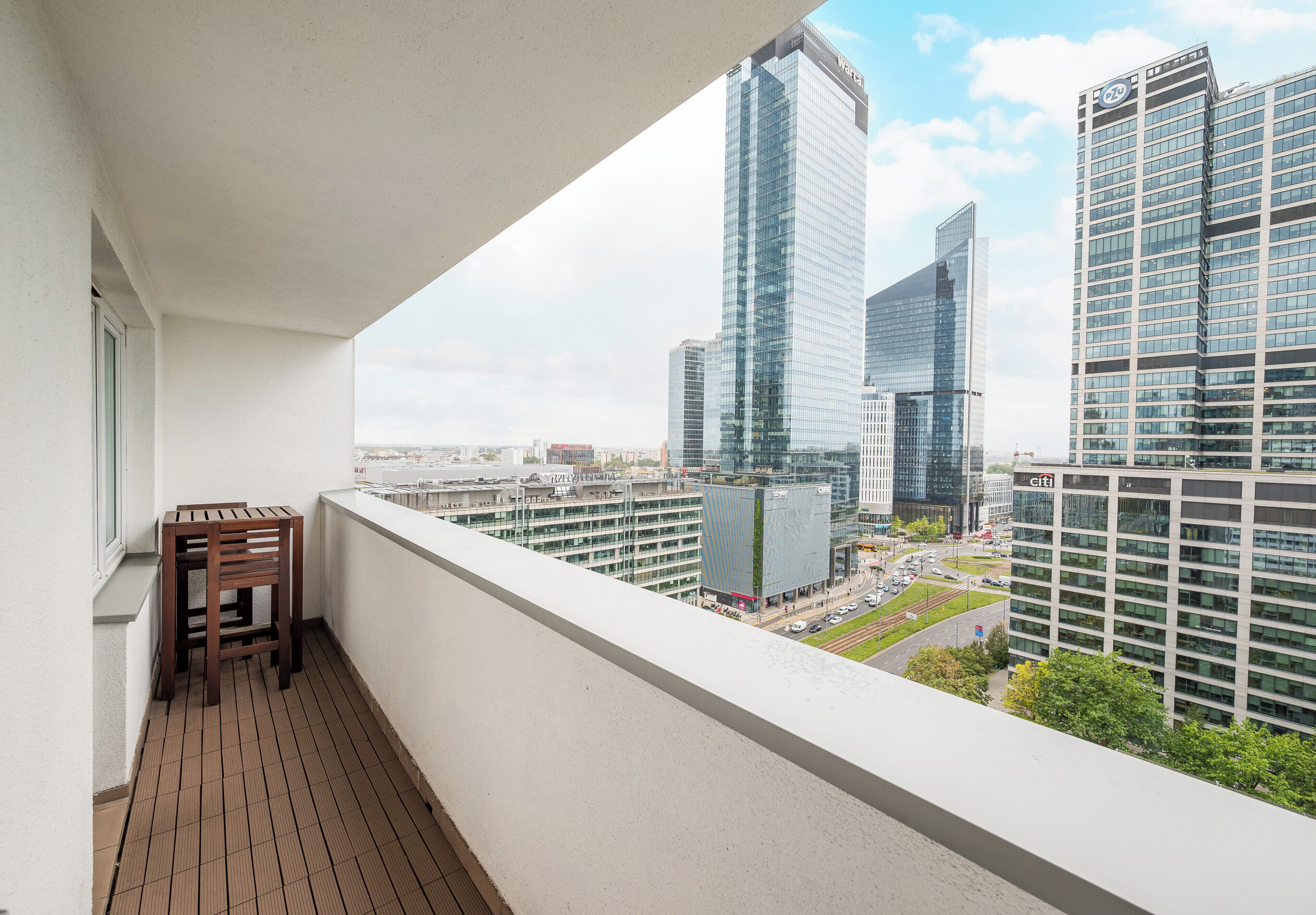 A balcony with a view of modern high-rise buildings, a wooden chair, and a tiled floor.