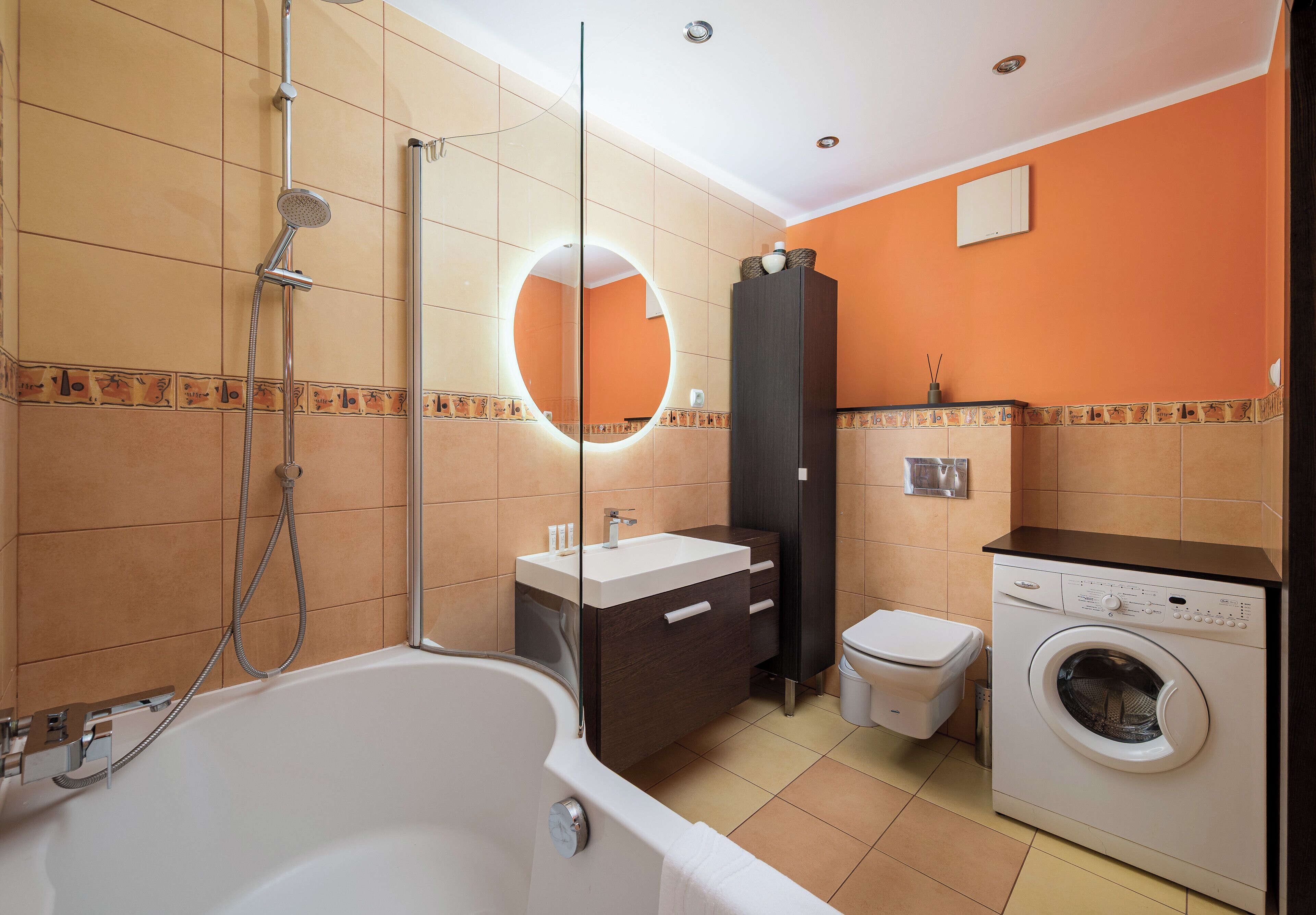 A bathroom with a bathtub, a toilet, a washing machine, and a mirror with a light.